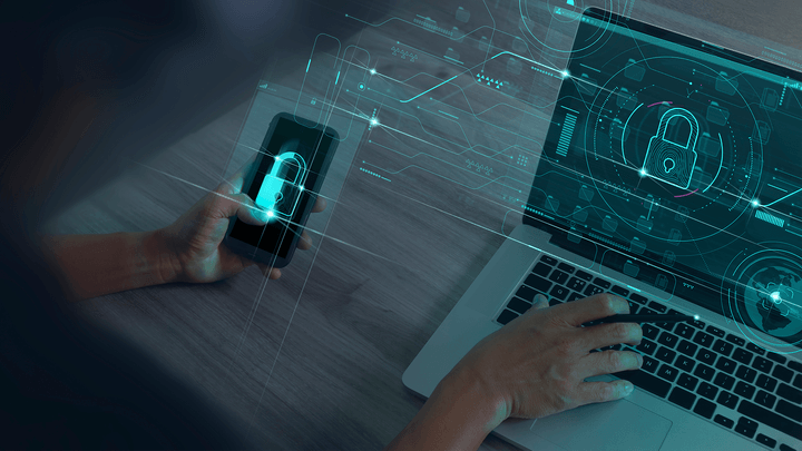 hand holding iphone and another hand typing on open laptop with cybersecurity lock symbols